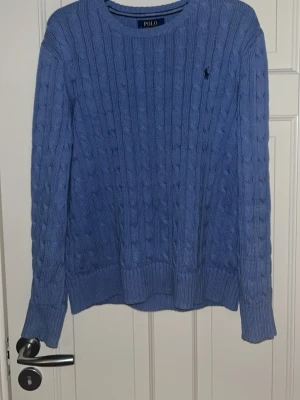 Blå kabelstickad tröja från Polo Ralph Lauren - Kabelstickad blå tröja från Polo Ralph Lauren med klassisk rund halsringning och broderad logga på bröstet. Tröjan har långa ärmar och ribbade muddar vid ärmslut och nederkant. Perfekt för dig som gillar preppy stil.