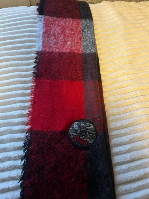 Rutig ullväst med Dio-märke - Säljer en rutig ullväst i mörkröda, svarta och grå toner. Västen har ett stort, runt Dio-tygmärke på framsidan med ett coolt motiv och röda detaljer. Materialet är mjukt och värmande ull, perfekt för dig som vill sticka ut med en unik detalj.