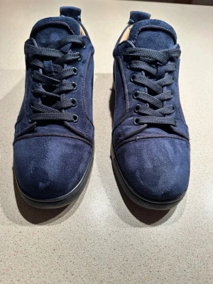 Marinblå mockasneakers från Louboutin - Säljer ett par marinblå sneakers från Christian Louboutin i mjuk mocka med klassisk röd sula och broderad logga på hälen. Skorna har rund tå, svarta snören och svart sula. Insidan är ljusbeige och designen är stilren men ändå lyxig.