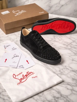 Christian Louboutin svarta sneakers med nitar - Säljer ett par svarta sneakers från Christian Louboutin i mocka med ikoniska röda sulor och svarta nitar över tån. Skorna har snörning och rund tå, samt exklusiv känsla med lyxiga detaljer. Perfekta för dig som vill sticka ut med edgy design.