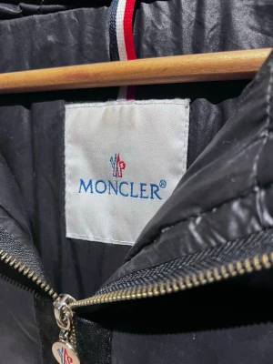 Svart Moncler pufferjacka med päls - Säljer en svart pufferjacka från Moncler med fluffig päls på huvan och guldfärgad logga på ärmen. Jackan har quiltad design, dragkedja framtill och klassisk Moncler-detalj på dragkedjan. Perfekt för kalla dagar och riktigt snygg till vintern. Det är en dam modell. 