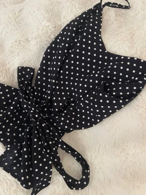 Svart prickig topp med knyt - Supersöt svart topp med vita prickar och knyt i midjan. Toppen har smala axelband och man knyter den så mycket man vill där bak. Perfekt för dig som gillar retrovibbar och vill sticka ut lite extra.