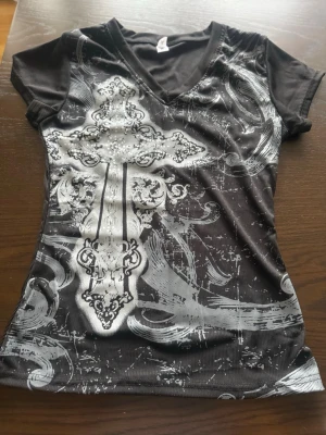 Svart t-shirt med silverkors - Snygg svart t-shirt med stort silvrigt kors och detaljerat mönster framtill. T-shirten har v-ringning och korta ärmar. Materialet känns stretchigt och följsamt, perfekt för dig som gillar statement-plagg med edge.