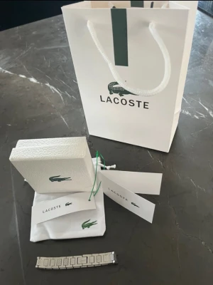 Snygg silverfärgad Lacoste länkarmband - Stilrent armband från Lacoste i silverfärgad metall med klassisk länkdesign och ingraverad krokodil på spännet. Kommer med original ask, påse och tags. Perfekt accessoar för dig som gillar en clean och modern look.