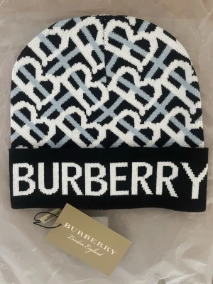 Burberry mössa - Snygg mössa från Burberry med svart, vit och ljusblått mönster och stor logga framtill. Klassisk ribbad kant och stickad i mjukt material. Perfekt accessoar för att lyfta din vinterstil.