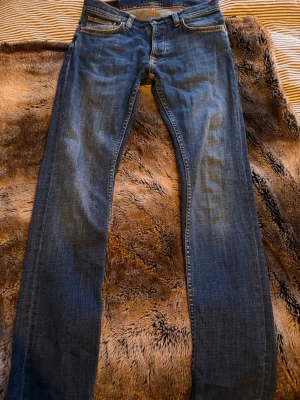 Blå jeans från Nudie Jeans - Säljer ett par klassiska blå jeans från Nudie Jeans med snygga kontrastsömmar och ikonisk broderad bakficka. Modellen har normal passform och raka ben, tillverkade i slitstarkt bomullstyg. Perfekta för dig som gillar tidlös och enkel stil.