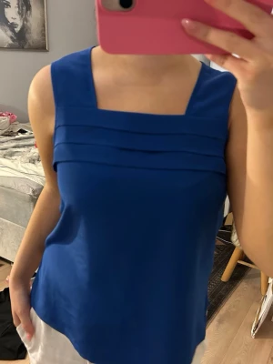 Kornblå ärmlös linnetopp - Snygg kornblå linnetopp med breda axelband och fyrkantig ringning. Toppen har dekorativa veck framtill och är tillverkad i ett mjukt, lätt material som känns skönt mot huden. Perfekt för varma dagar och enkel att styla till olika outfits.