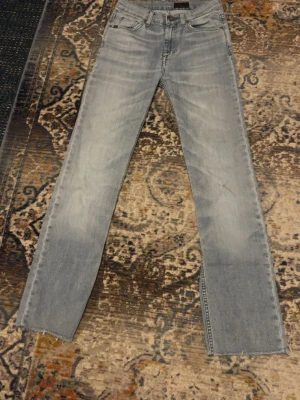 Ljusblå jeans från Tiger of Sweden - Säljer ett par ljusblå jeans från Tiger of Sweden med klassisk femficksdesign och raka ben. Jeansen har en snygg tvättad look och detaljer som läderpatch bak i midjan. Perfekta till sneakers och riktigt sköna att bära.