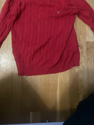 Röd kabelstickad tröja från GANT - Säljer en röd kabelstickad tröja från GANT i premium bomull. Tröjan har rund hals, diskret logga på bröstet och ribbade muddar. Perfekt för dig som vill ha en klassisk och stilren look.