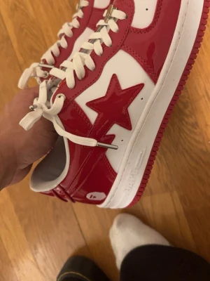BAPESTA sneakers röd/vit - Säljer ett par BAPESTA sneakers i rött och vitt med glansig finish och ikonisk stjärndetalj på sidan. Skorna har vita snören, perforerad tå och klassisk låg siluett. Sulan är vit med röd undersida och BAPE-logga bakpå hälen. Perfekta för dig som vill sticka ut.