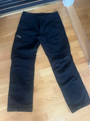 Svarta täckbyxor från Lundhags strl 52 - Säljer ett par svarta täckbyxor från Lundhags i storlek 52. Byxorna har flera praktiska fickor på benen och förstärkta partier vid knäna. Perfekta för utomhusaktiviteter och äventyr. Klassisk design med justerbara benslut.