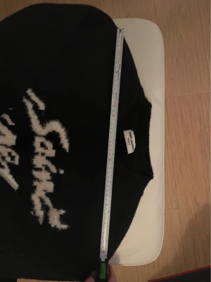 Svart stickad Saint Laurent tröja - Säljer en svart stickad tröja från Saint Laurent med vit, fluffig text på bröstet. Tröjan har klassisk rund halsringning och är i mjukt material som känns lyxigt. Perfekt för dig som vill ha något stilrent men ändå med en twist.