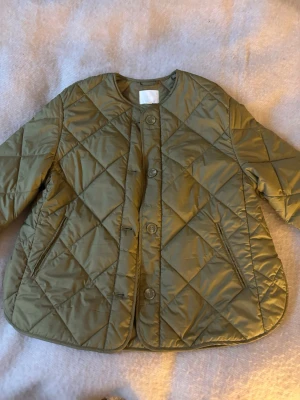 Olivgrön pufferjacka H&M - Säljer en olivgrön pufferjacka med quiltat mönster och rund hals i storlek S. Jackan har knäppning med stora knappar framtill och två fickor på sidorna. Den har en loose passform och är perfekt för höst, vår och vinter💞