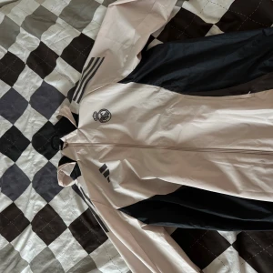 Beige vindjacka Real Madrid Adidas L - Snygg beige och svart vindjacka från Adidas med Real Madrid-logga på bröstet. Jackan har huva, hel dragkedja och klassiska Adidas-ränder på axeln. Tillverkad i lätt polyester, perfekt för blåsiga dagar. Sportig look med meshfoder och elastiska muddar. Säljes för den är för stor