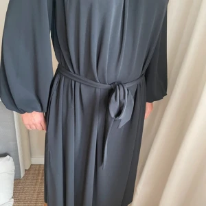 Mörkgrå klänning från Day Birger et Mikkelsen - Snygg mörkgrå långklänning från Day Birger et Mikkelsen med långa vida ärmar och knytskärp i midjan. Klänningen har en avslappnad passform och är tillverkad i ett mjukt, följsamt material som ger ett elegant fall.