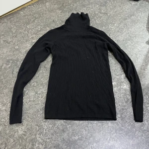 Svart ribbad polotröja XS - Svart ribbad polotröja från C i storlek XS. Tröjan har lång ärm och hög polokrage, perfekt för lager-på-lager. Den är figurnära och tillverkad i ett stretchigt, mjukt material som sitter snyggt på kroppen.