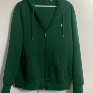 Grön hoodie från Polo Ralph Lauren - Mörkgrön hoodie från  Ralph Lauren med dragkedja, huva och snörning. Perfekt för chill dagar eller när du vill vara extra bekväm. Flexibel med priset