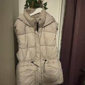 Beige dunväst med huva från H&M - Beige quiltad dunväst från H&M i storlek L. Västen har en stor huva, dragsko i midjan och praktiska fickor framtill. Perfekt för lager-på-lager och chill höst- eller vinterstil.