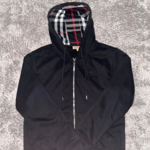 Svart Burberry hoodie med dragkedja - Svart hoodie från Burberry med klassiskt rutigt foder i huvan och broderad logga på bröstet. Tröjan har hel dragkedja, snörning i huvan och två fickor framtill. Perfekt för dig som gillar stilrena och exklusiva plagg.