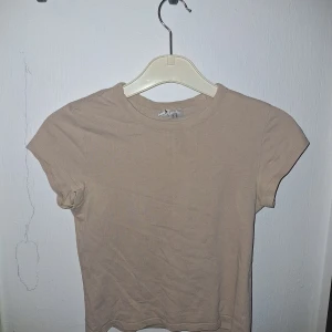 Beige t-shirt - En basic beige t-shirt från H&M Divided i storlek XS. Modellen är kortärmad med rund halsringning och har en enkel, clean look. Tillverkad i mjuk bomull som är skön mot huden. Perfekt till jeans eller kjol för en chill vibe.