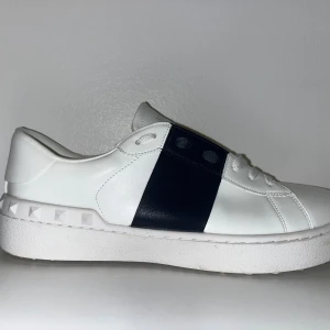 Valentino Garavani Open sneakers vit/mörkblå - Säljer ett par snygga Valentino Garavani Open sneakers i vitt skinn med bred svart rem över mitten. Skorna har coola nitar runt hälen och klassisk rund tå. Perfekta för dig som gillar stilrena men ändå unika sneakers.