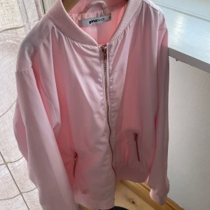 Ljusrosa bomberjacka från Gina Tricot - Snygg ljusrosa bomberjacka från Gina Tricot med dragkedja framtill och två fickor med dragkedja. Jackan har ribbade muddar vid hals, ärmslut och nederkant. Perfekt för dig som vill ha en stilren och trendig look.