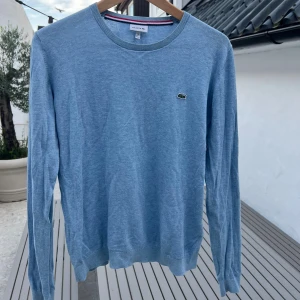 Ljusblå stickad tröja från Lacoste - Säljer en ljusblå stickad tröja från Lacoste med klassisk rund halsringning och den ikoniska krokodilloggan broderad på bröstet. Tröjan har långa ärmar och ribbade muddar för en snygg passform. Perfekt för dig som gillar stilren och enkel design.