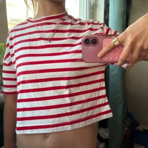 Randig croppad t-shirt i bomull - Säljer en croppad t-shirt med röda och vita horisontella ränder. Toppen har rund hals och korta ärmar, perfekt för dig som gillar en chill och fräsch stil. Materialet är mjuk bomull och passformen är normal. Enkel att matcha med jeans eller shorts.