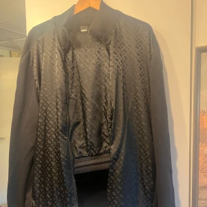 Svart bomberjacka från BOSS - Snygg svart tracksuit från BOSS med diskret mönster över hela framsidan. Jackan har ribbad krage och muddar samt dragkedja framtill. Materialet är blankt och känns lyxigt, perfekt för dig som vill ha en stilren och modern look. Använd 1 gång 
