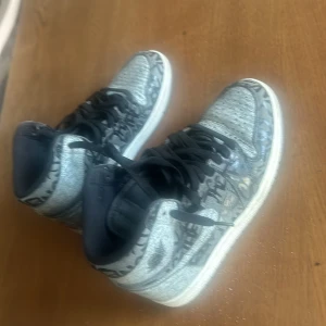 Nike Air Jordan 1 grå/svarta sneakers - Säljer ett par Nike Air Jordan 1 sneakers i grått och svart med coolt grafiskt mönster. Skorna har högt skaft, svarta snören och vit sula. Perfekta för dig som vill sticka ut med en unik streetstyle-look.