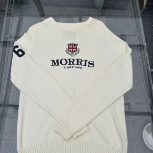 Morris stickad tröja – stl L - Supersnygg och tidlös tröja från Morris i offwhite/cream. Klassisk modell med broderad Morris-logga, “Since 1968”-text och Union Jack-emblem framtill. Nummer 6 på axeln ger en sportig touch.  Tillverkad i 100% bomull, så den är både mjuk och håller formen bra. Passar perfekt till jeans, chinos eller över en skjorta för en clean, preppy look.  ✨ Storlek: Large ✨ Material: 100% bomull ✨ Färg: Offwhite / Cream ✨ Skick: Mycket bra – inga fläckar eller defekter  En riktig klassiker från Morris – stil