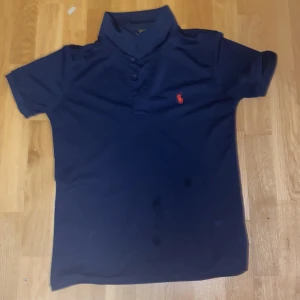 Marinblå pikétröja från Polo Ralph Lauren - Klassisk marinblå pikétröja från Polo Ralph Lauren med röd broderad logga på bröstet. Tröjan har korta ärmar, krage och knappar framtill. Perfekt för en clean och sportig look.