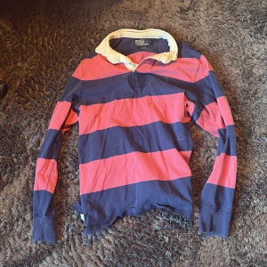 Randig pikétröja Polo Ralph Lauren M - Klassisk långärmad pikétröja från Polo Ralph Lauren med breda marinblå och röda ränder, vit krage och broderad grön logga på bröstet. Tröjan är i bomull och har en relaxed fit med sportig känsla. Perfekt för dig som gillar preppy stil.