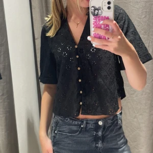 Svart blus med broderade detaljer - Svart blus med kort ärm och v-ringning. Blusen har broderade hålmönster i blommig stil och knäpps framtill med små knappar. Croppad passform som ger en trendig look. Perfekt att matcha med jeans eller kjol.