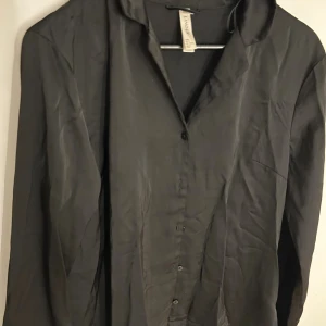 Svart silkesliknande skjorta från Lindex  - Stilren svart skjorta från Lindex i silkesliknande material. Skjortan har klassisk krage, knäppning framtill och långa ärmar. Perfekt för dig som gillar en clean och enkel look med lite glans i tyget.