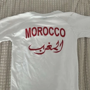Marocko vit fotbollströja Puma - Säljer en officiell Marocko fotbollströja från Puma i vitt med röda detaljer. Tröjan har korta ärmar, V-ringning och tunna grå linjer över axlarna. På bröstet finns Marockos landslagsemblem och Puma-logga. Materialet är lätt och andas, perfekt för match eller träning.