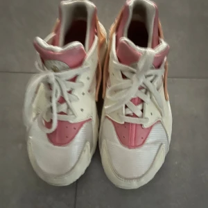 Nike sneakers i vitt och rosa - Nike sneakers i vitt, rosa och persikofärgat med klassisk snörning och rund tå. Skorna har en cool design med bred hälrem där det står NIKE och detaljer i syntetmaterial. Perfekta för dig som vill sticka ut med färg på fötterna.