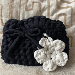 Svart handväska med virkad blomma - Unik svart handväska i grovt virkat tyg med en stor vit virkad blomma som detalj. Väskan har en söt knytrosett framtill och är perfekt för dig som vill sticka ut med en personlig accessoar.