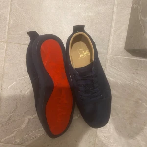 Mörkblå Christian Louboutin sneakers - Säljer ett par mörkblå sneakers från Christian Louboutin i mocka med klassisk röd sula. Skorna har rund tå, platt sula och snörning. Insidan är beige och märkt med Louboutins logga. Perfekta för dig som vill sticka ut med exklusiva detaljer.