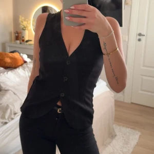 Svart kostymväst från H&M - Klassisk svart kostymväst från H&M i figurnära modell med tre knappar framtill och v-ringad hals. Västen har en stilren look och är tillverkad i ett slätt tyg som ger en dressad känsla. Perfekt att styla över en t-shirt eller skjorta.