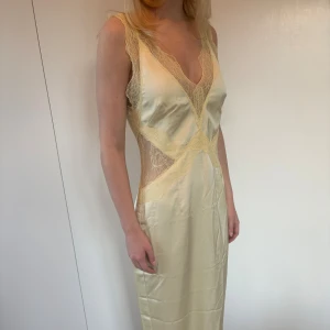 Gul långklänning i satin - Ljusgul långklänning i satin material med v-ringning och spetsdetaljer vid byst och midja. Ärmlös med figurnära passform och transparent spets i sidorna. Perfekt för sommaren eller exempelvis studentbalen! Aldrig använd, endast provad