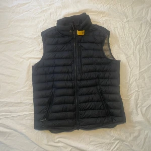 Svart dunväst från Parajumpers M - Svart dunväst från Parajumpers i storlek M. Västen är quiltad med horisontella sömmar, har hög krage, dragkedja framtill och två fickor med dragkedja. Klassisk Parajumpers-logga i nacken. Den har ett litet litet hål på nedre rygg, och ett litet hål på innersidan samt en liten fläck. (Se bilder) Fläcken och hålet syns ingenting på utsidan. 