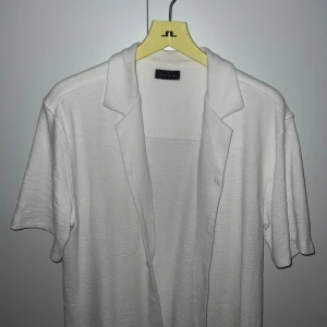 Vit kortärmad skjorta från Brand Island - Säljer en stilren vit skjorta från Brand Island med korta ärmar och klassisk krage. Skjortan har knappar framtill och är gjord i ett mjukt, strukturerat material som känns luftigt. Perfekt för dig som gillar en clean och enkel look.