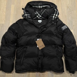 Svart pufferjacka från Burberry - Säljer en svart pufferjacka från Burberry med klassiskt rutigt foder i huvan. Jackan har dragkedja och knappar framtill, två sidofickor med dragkedja och Burberry-logga på ärmen. Perfekt för kalla dagar och riktigt snygg streetstil.