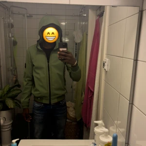 Grön vindjacka från Stone Island - Säljer en grön vindjacka från Stone Island med huva och svart dragkedja. Jackan har den klassiska loggan på vänster ärm och är tillverkad i 100% polyester. Snygg och stilren modell med normal passform och långa ärmar.