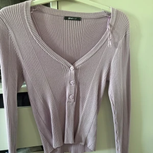 Ljuslila ribbad topp från Gina Tricot - Superfin ljuslila ribbad topp från Gina Tricot med v-ringning och knappar framtill. Toppen har lång ärm och croppad passform, perfekt att styla med höga jeans eller kjol. Enkel men ändå trendig med sitt ribbade material och snygga detaljer.
