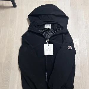 Svart Moncler dunjacka med huva - Svart dunjacka från Moncler med huva och dragkedja framtill. Jackan har klassisk Moncler-logga på ärmen och innerfoder med serietidningsmönster. Tillverkad i polyester och dun, perfekt för kyliga dagar. Snygg och stilren design som passar till det mesta.