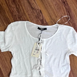 Vit ribbad knytblus från Bik Bok - Superfin vit ribbad blus från Bik Bok i ekologisk bomull. Toppen har korta ärmar, rund halsringning och snygga knytningar framtill som ger en trendig look. Perfekt att styla med jeans eller kjol för en fräsch vibe.