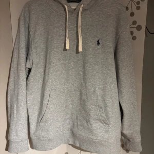 Grå hoodie från Polo Ralph Lauren - Säljer en grå hoodie från Polo Ralph Lauren i storlek M. Tröjan har huva med vita snören, känguruficka och det klassiska blå Polo-logot på bröstet. Mjuk bomullskvalitet och ribbade muddar vid ärmslut och nederkant. Perfekt för chill dagar.