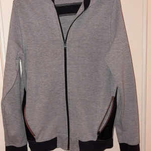 Grå hoodie från Hugo Boss med dragkedja - Snygg grå hoodie från Hugo Boss med svart logga på ryggen. Tröjan har hel dragkedja framtill, svarta detaljer vid fickorna och muddar i mörkare nyans. Perfekt för en chill och stilren look.